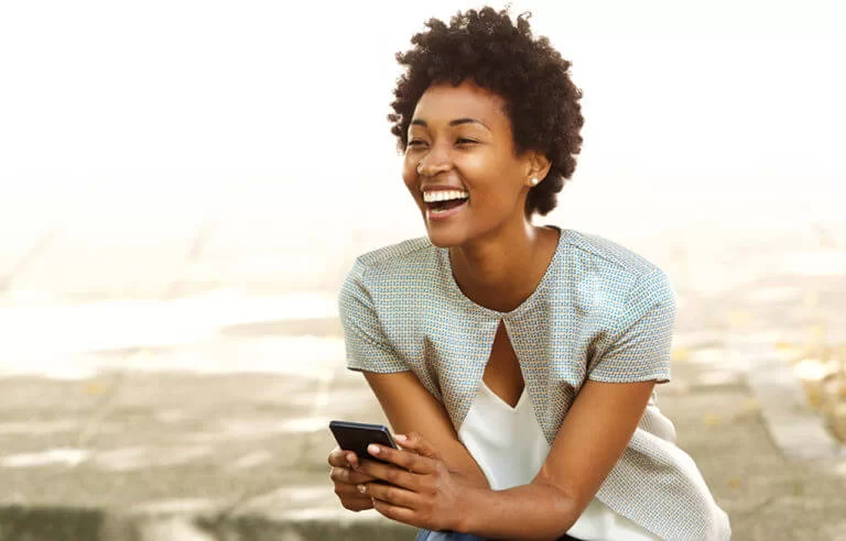 Mulher negra jovem sorrindo com um celular nas mãos demonstrando alegria e sem stress