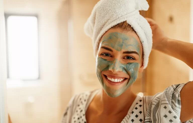 Mulher sorrindo com máscara de tratamento verde no rosto e toalha de banho no cabelo