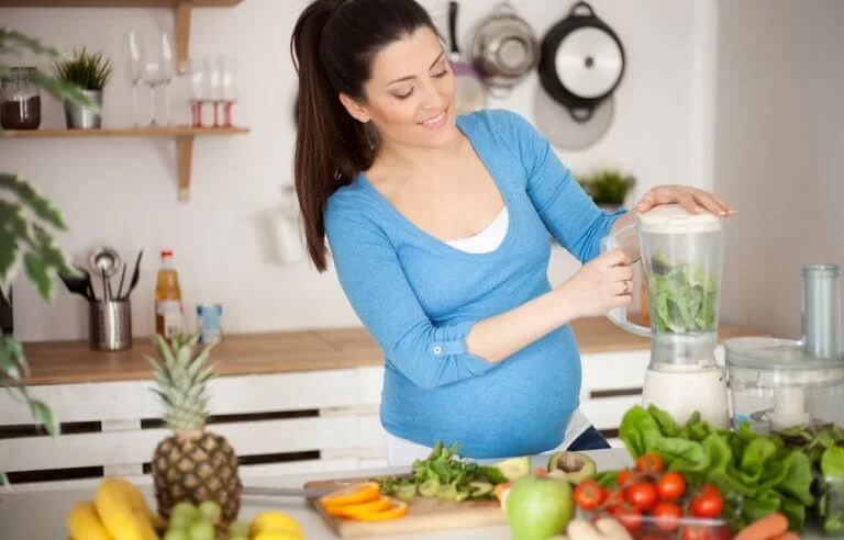 Grávida batendo verdura no liquidificador e com frutas e legumes na cozinha