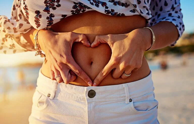 Mulher fazendo coração com os dedos em cima de sua própria barriga com o umbigo no centro