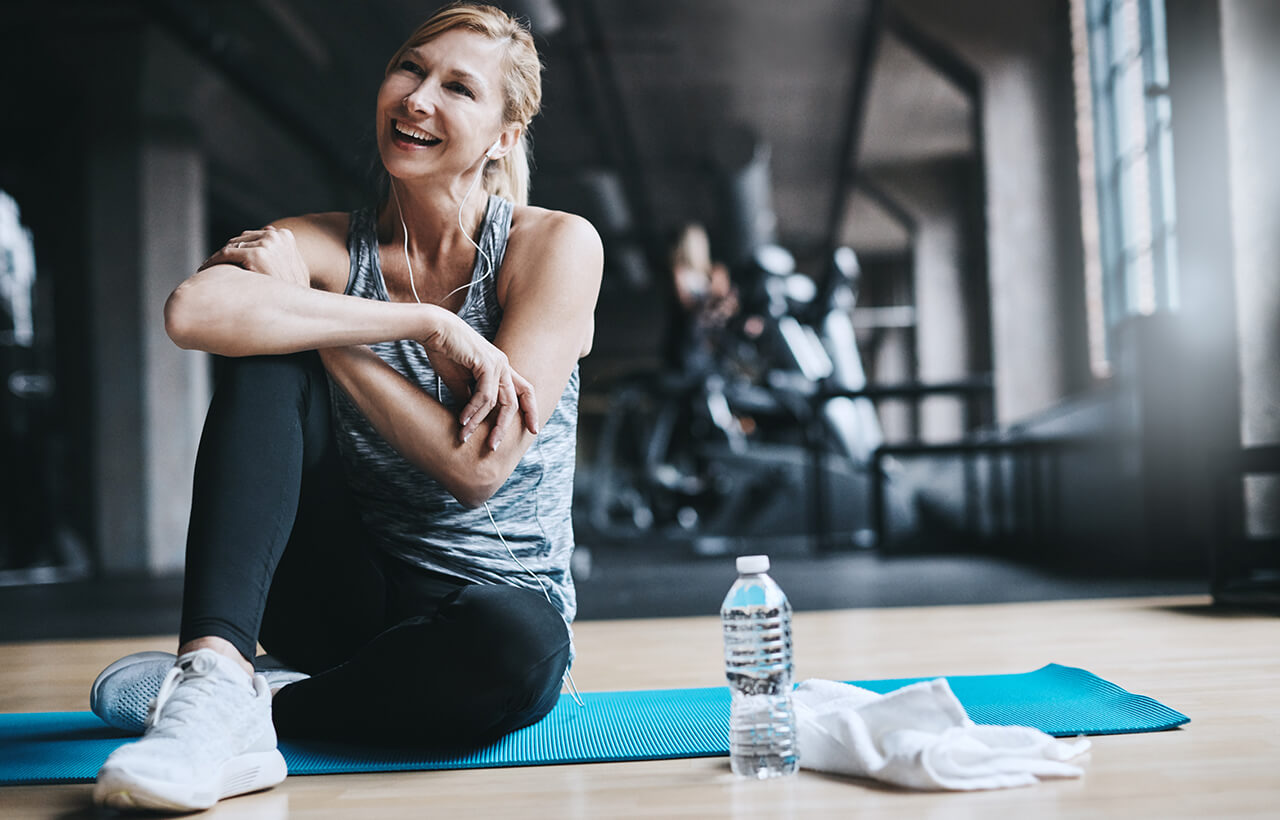 Mulher loira sorrindo na academia sentada em tapete de yoga e ao fundo mulher malhando