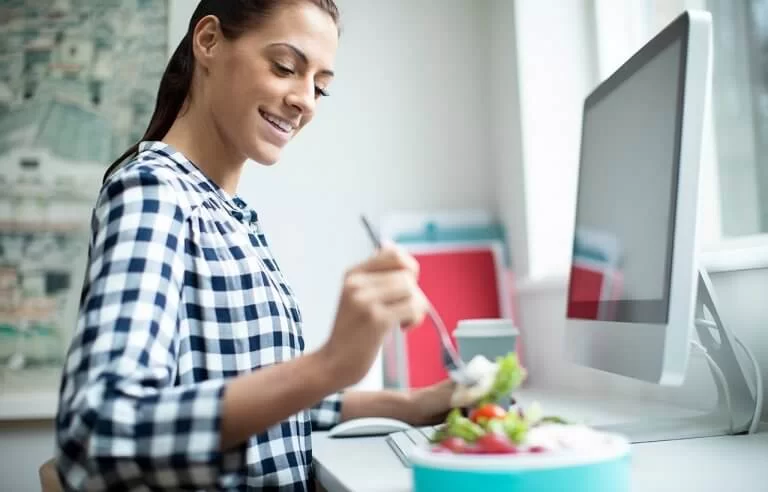 Mulher de roupa xadrez sorrindo enquanto come uma salada em frente ao computador