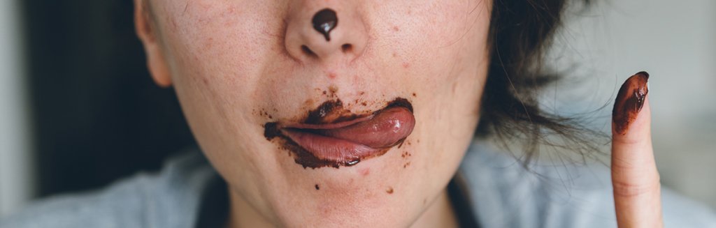 Pessoa com os dedos, boca e nariz sujos de chocolate se lambuzando.