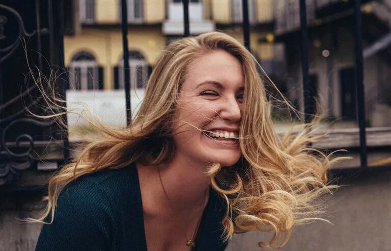 Mulher loira com cabelos ao vento com um grande sorriso no rosto no quintal de casa