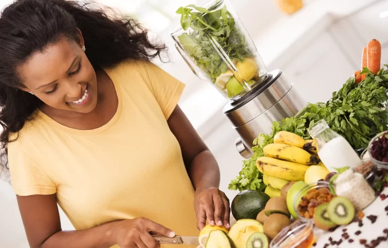 Mulher negra sorrindo enquanto corta laranja para preparar uma bebida saudável como suco verde ou vitamina