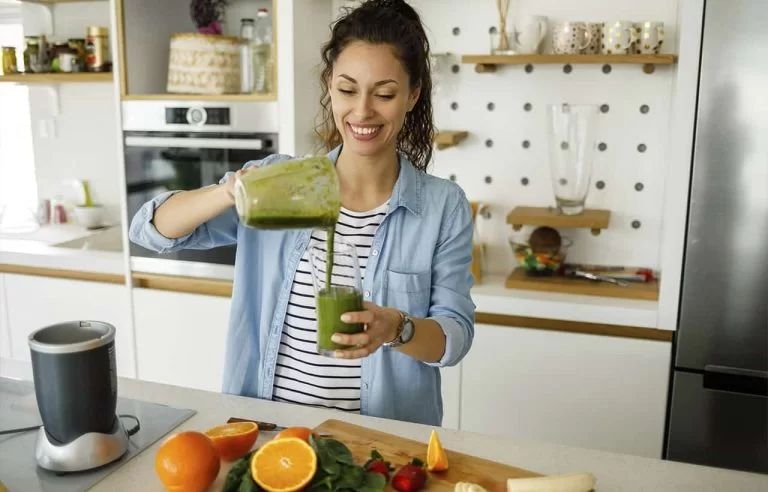 Mulher na cozinha sorrindo prestes a tomar um suco verde com frutas e verduras perto dela