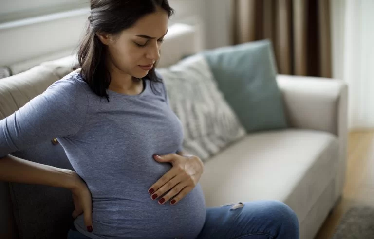 Mulher grávida com expressão de dor sentada no sofá com as mãos na barriga