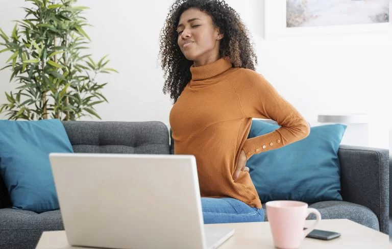 Mulher negra sentindo dor nas costas enquanto trabalha no sofá durante home office