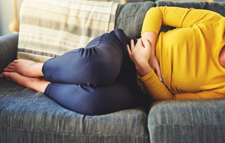Mulher deitada no sofá com as duas mãos na barriga simbolizando dor na barriga ou cólica