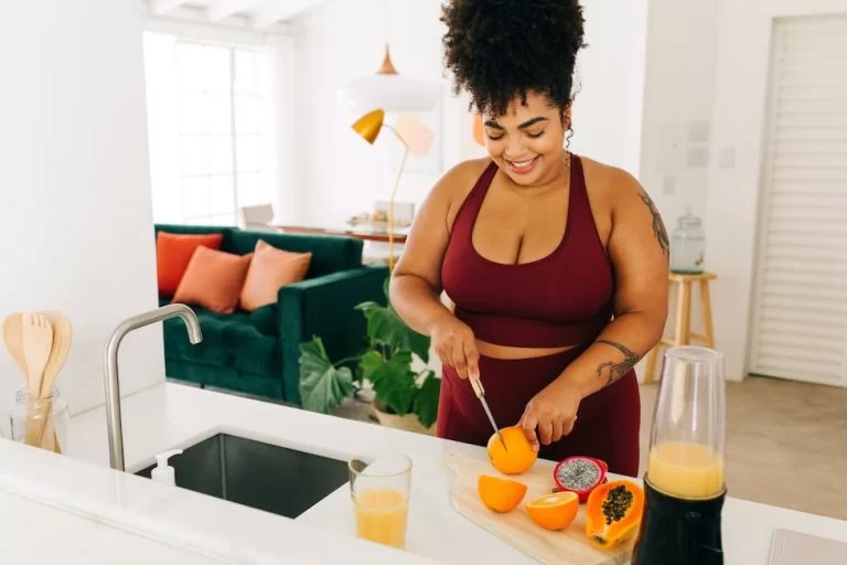 Mulher feliz cortando laranja e ao lado há mamão cortado e liquidificador com suco amarelo