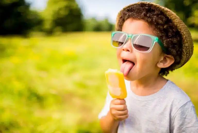 Menino de óculos de sol e chapéu tomando picolé amarelo na natureza: vida saudável e feliz