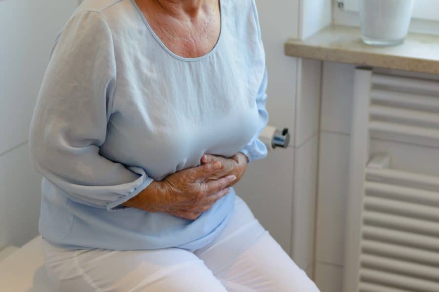 Idosa de blusa azul e calça branca com as mãos na barriga sofrendo com constipação
