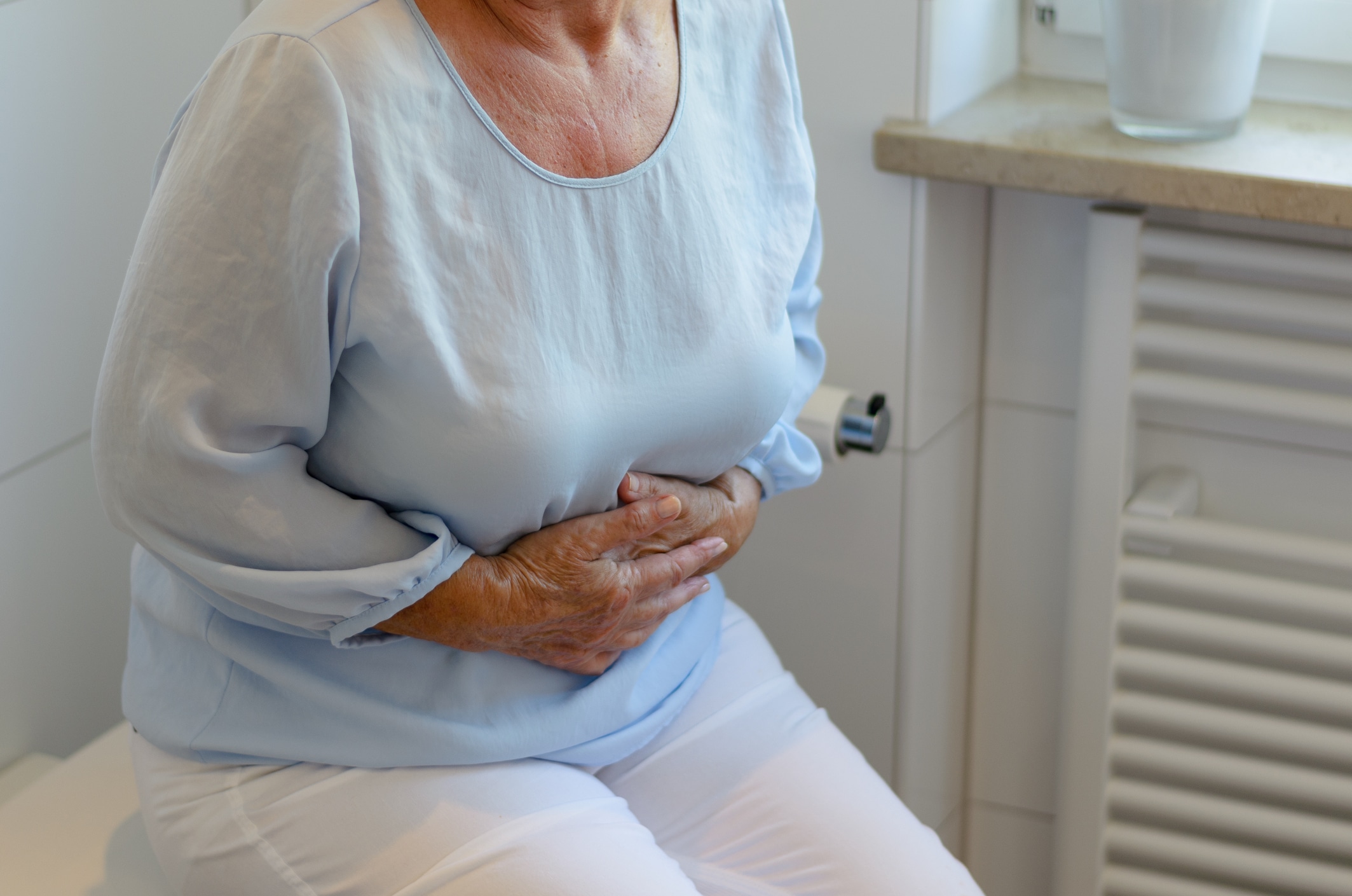Idosa de blusa azul e calça branca com as mãos na barriga sofrendo com constipação
